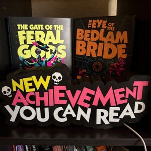 May include: Two books titled "The Gate of the Feral Gods" and "The Eye of the Bedlam Bride" are displayed on a shelf. A black sign with the words "NEW ACHIEVEMENT YOU CAN READ" in white, pink, and yellow is in front of the books. The sign has two skull graphics.