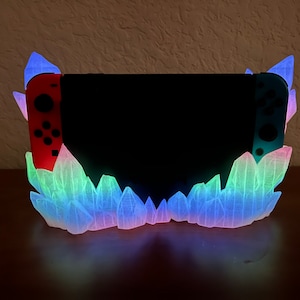 May include: A Nintendo Switch console is displayed in a crystal-shaped stand. The stand is made of clear plastic and glows in a rainbow of colors, including red, orange, yellow, green, blue, and purple.