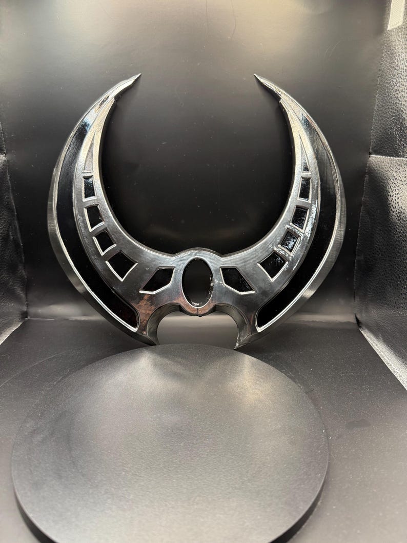 May include: A silver-colored metal crescent moon-shaped object with intricate details and a black center. The object is on a black circular base.