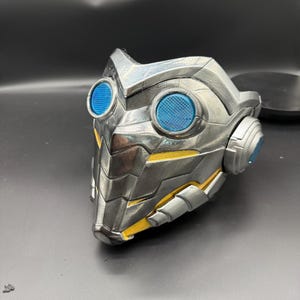 May include: A silver and yellow 3D printed mask with blue lenses. The mask has a futuristic design with a textured surface.