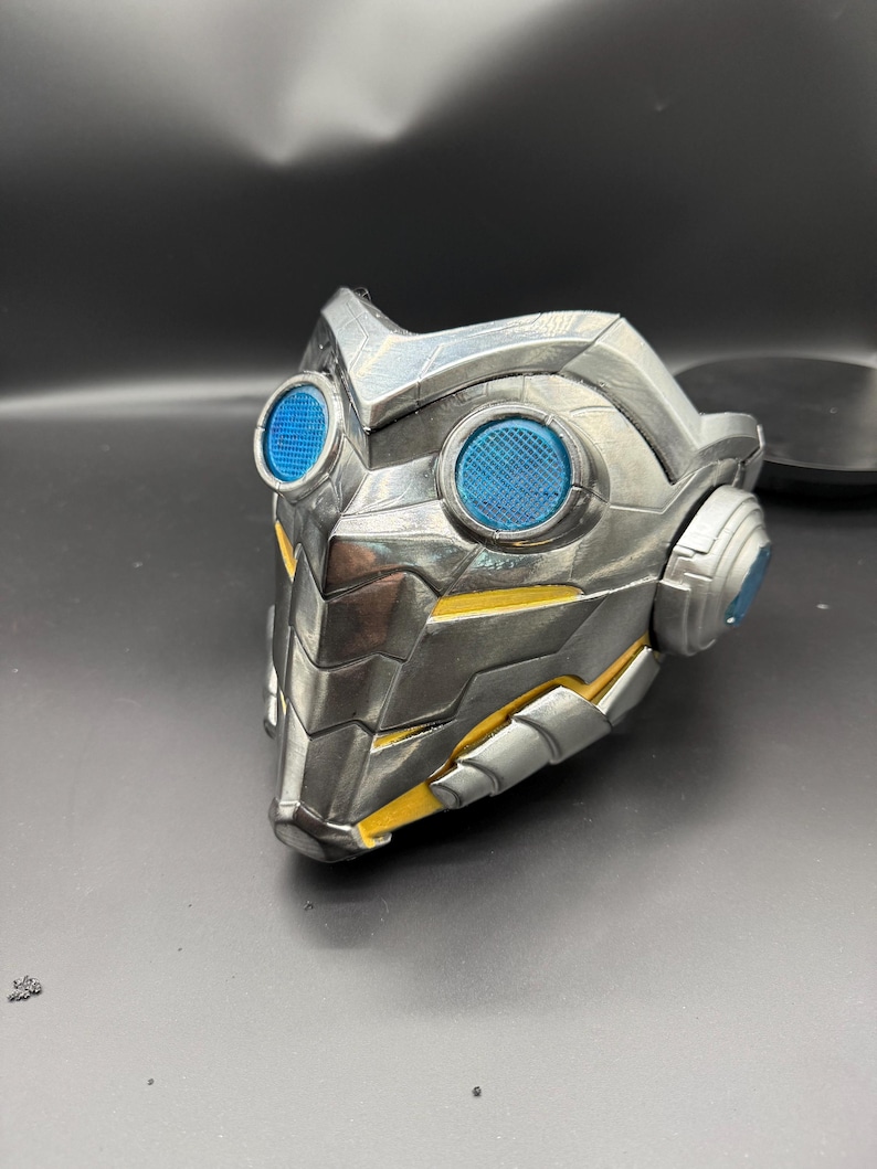 May include: A silver and yellow 3D printed mask with blue lenses. The mask has a futuristic design with a textured surface.