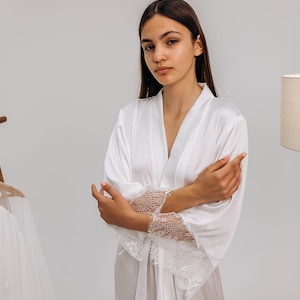 Puede incluir: Una bata de satén blanca con ribetes de encaje en las mangas. La bata tiene un cinturón de lazo en la cintura.