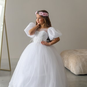 May include: A young girl wearing a white tulle dress with a beaded bodice and puff sleeves. She is wearing a pink flower crown and has long brown hair.