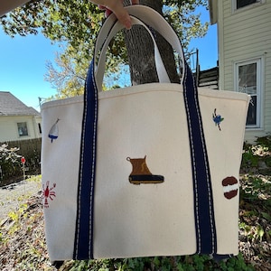 May include: A cream-colored canvas tote bag with two navy blue handles and navy blue vertical stripes. The bag features embroidered motifs including a brown and black duck boot, a red lobster, a blue fishing buoy, a blueberry, and a brown cookie.