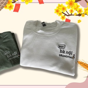 May include: Two sweatshirts with the text "Best! Bà nội Grandma" in a speech bubble. The sweatshirts are green and white. The background is a pink and white floral design.