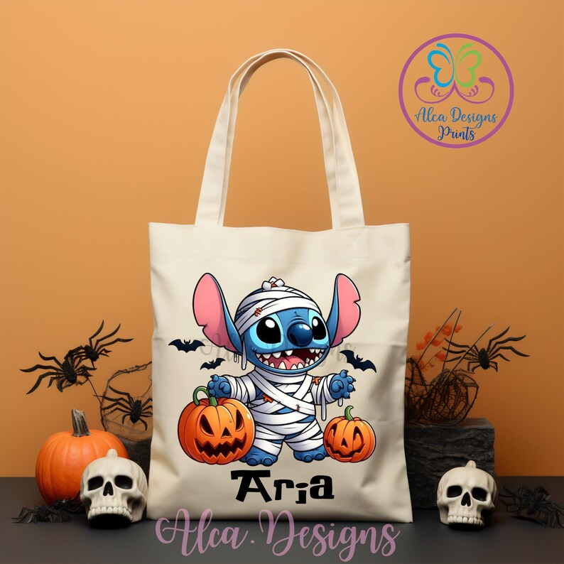 May include: A cream-colored tote bag featuring a cartoon Stitch character dressed as a mummy, holding two orange jack-o'-lanterns. The bag has black bats and the name "Aria" printed on it, with the text "Alca Designs Prints".