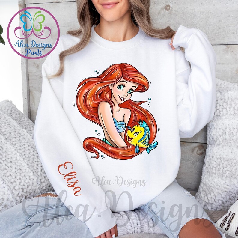 Op de afbeelding: Witte trui met een roodharige zeemeermin en een gele vis die op de voorkant zijn gedrukt. De tekst "Elisa" is gedrukt op de linkermouw.