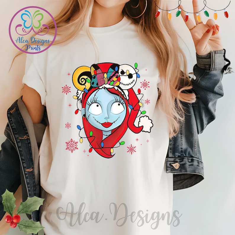 Op de afbeelding: Wit T-shirt met een kleurrijke illustratie van Sally uit The Nightmare Before Christmas die een kerstmanmuts draagt met kerstverlichting. De illustratie is omringd door sneeuwvlokken en kerstverlichting.
