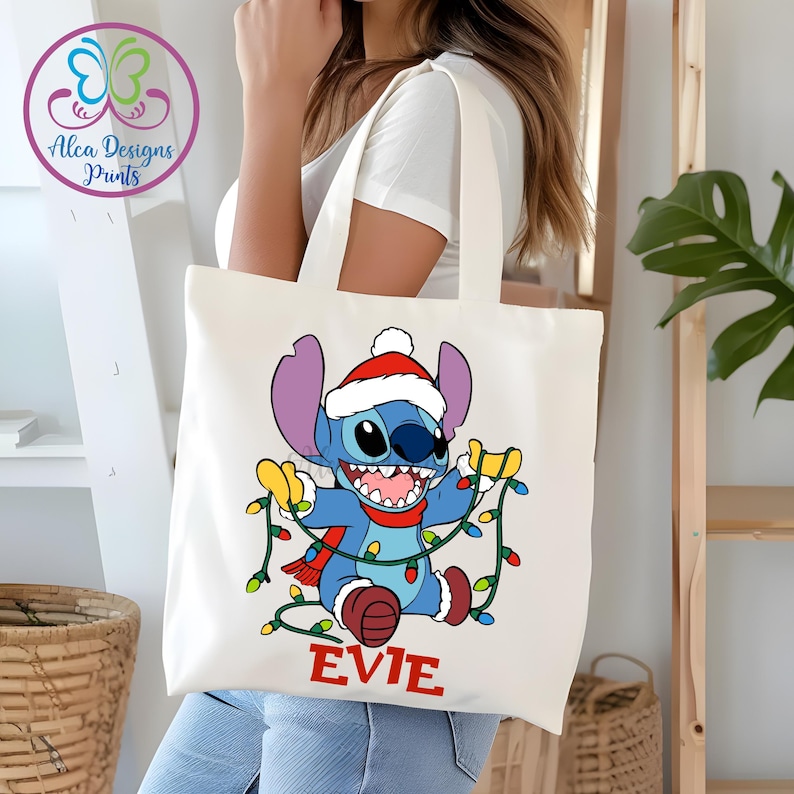 Puede incluir: Un bolso tote blanco con un personaje de dibujos animados que lleva un gorro y una bufanda de Pap&aacute; Noel, sosteniendo luces navide&ntilde;as. El personaje es azul con orejas grandes y una amplia sonrisa. El nombre "EVIE" est&aacute; impreso en rojo en la parte inferior.