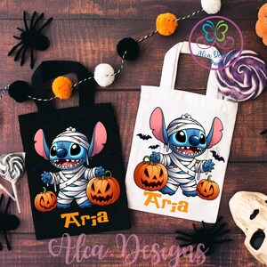 May include: Two Halloween-themed tote bags, one black and one white, featuring a cartoon character dressed as a mummy holding pumpkins. The name "Aria" is printed in orange. Decorative spiders and a pom-pom garland are in the background.