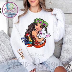 May include: White sweatshirt featuring a colourful cartoon image of a girl with a flower crown, a pig, and a rooster. The text "Elisa" is on the sleeve of the sweatshirt.