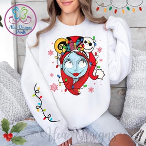 Op de afbeelding: Witte trui met een kleurrijke afbeelding van een personage uit The Nightmare Before Christmas dat een kerstmanmuts draagt en omringd is door kerstverlichting. Het personage heeft blauwe huid, zwart haar en rode lippen.