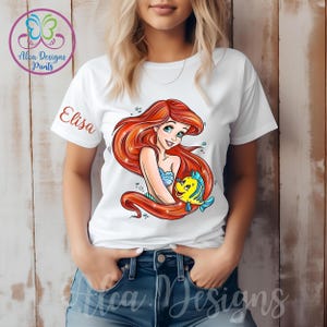Op de afbeelding: Wit T-shirt met een rode haren zeemeermin en een gele vis afbeelding. De tekst "Elisa" is op het T-shirt gedrukt.