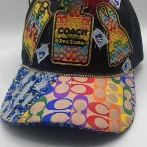 May include: Black baseball cap with a rainbow-colored brim and decorative patches. The patches include a perfume bottle design with the text "COACH PERFUME" and other colorful designs. The cap is embellished with blue and gold beads and colorful feathers.