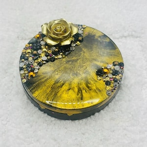 May include: A round, decorative box with a gold rose on top. The box has a black base and a gold and black marbled design. The top is embellished with gold, black, and iridescent rhinestones. A unique jewelry box or trinket holder.