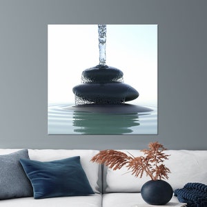 May include: A canvas print depicting a zen-like scene. Three dark gray stones are stacked, with water cascading over them into a pool of rippling water. The background is a soft, light blue, creating a sense of calm and tranquility.