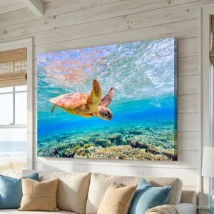 May include: A large canvas print of a sea turtle swimming underwater in a vibrant blue ocean. The artwork is displayed above a beige sofa in a coastal-themed living room with natural light and beach views.