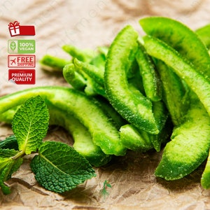 May include: A close-up of green candied citrus fruit slices on a brown paper background. The fruit is arranged in a pile, with some slices overlapping. There are also a few green mint leaves in the image.
