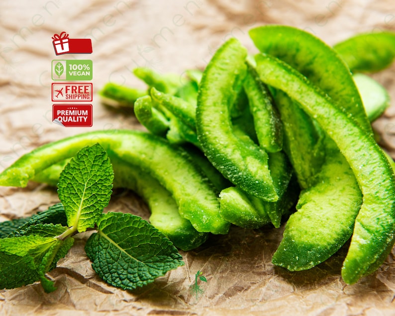 May include: A close-up of green candied citrus fruit slices on a brown paper background. The fruit is arranged in a pile, with some slices overlapping. There are also a few green mint leaves in the image.