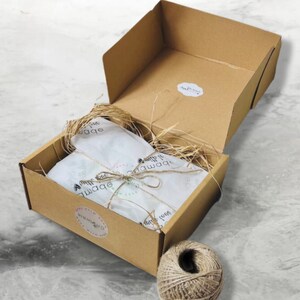 May include: A brown cardboard box with a white tissue paper lining and a brown twine bow. The box is open and the tissue paper is visible. The box is sitting on a white marble surface. There is a ball of twine next to the box.