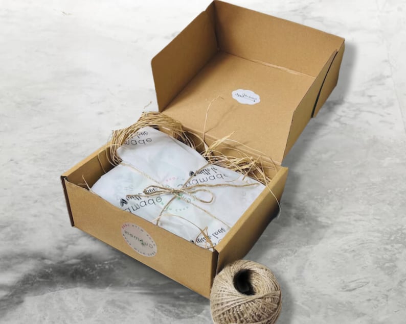 May include: A brown cardboard box with a white tissue paper lining and a brown twine bow. The box is open and the tissue paper is visible. The box is sitting on a white marble surface. There is a ball of twine next to the box.