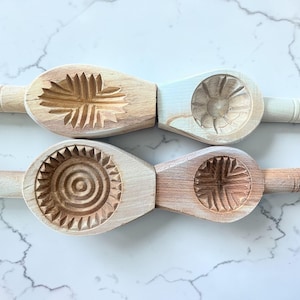 May include: Four wooden cookie molds with carved floral and geometric designs. Each mold has a handle and a distinct pattern, including a sunburst, a flower, and a cross-like design. The molds are made of light-colored wood.