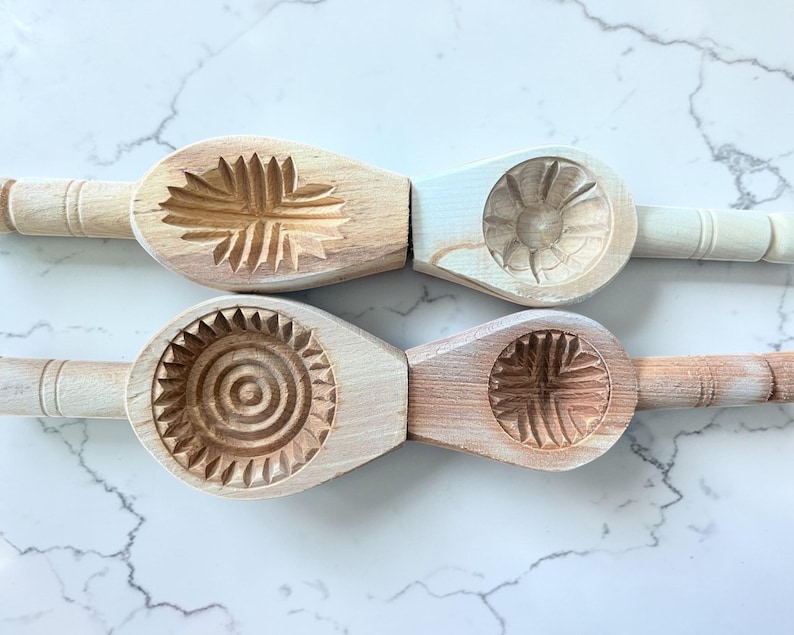 May include: Four wooden cookie molds with carved floral and geometric designs. Each mold has a handle and a distinct pattern, including a sunburst, a flower, and a cross-like design. The molds are made of light-colored wood.