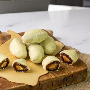 Puede incluir: Primer plano de una tabla de cortar de madera con dátiles cubiertos de chocolate blanco. Algunos de los dátiles están cortados por la mitad, revelando el relleno de dátiles marrones y una sola almendra en el interior.