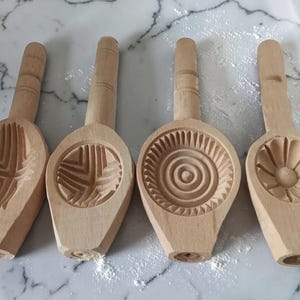 May include: Four wooden cookie molds with different carved designs. Each mold has a handle and a patterned impression area. The designs include geometric shapes and a floral pattern. The molds are made of light-colored wood.