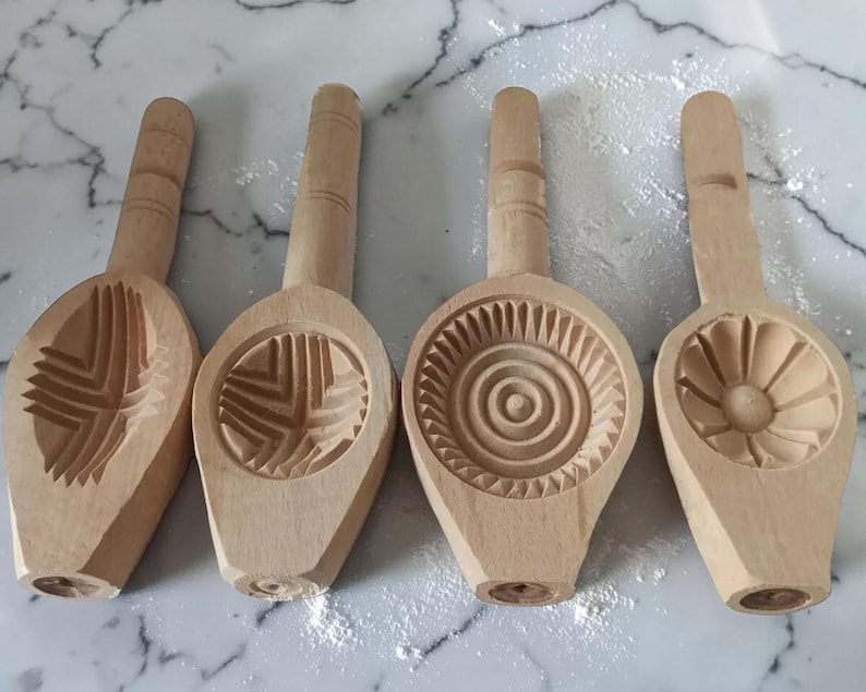 May include: Four wooden cookie molds with different carved designs. Each mold has a handle and a patterned impression area. The designs include geometric shapes and a floral pattern. The molds are made of light-colored wood.
