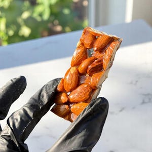 Op de afbeelding: Een close-up van een nougat reep met amandelen. De reep is lichtbruin van kleur en de amandelen zijn donkerbruin. De reep wordt vastgehouden in een zwarte handschoen.