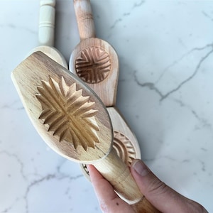 May include: Wooden cookie molds with carved designs. The molds have a handle and a detailed pattern on the base. One mold is held by a hand. The molds are made of light-colored wood and are arranged on a marble surface.