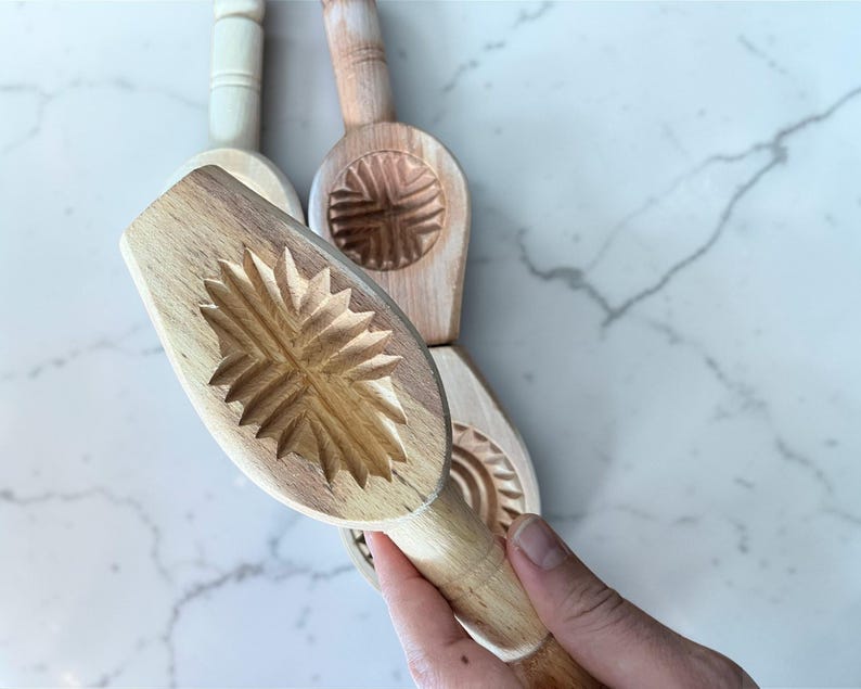 May include: Wooden cookie molds with carved designs. The molds have a handle and a detailed pattern on the base. One mold is held by a hand. The molds are made of light-colored wood and are arranged on a marble surface.