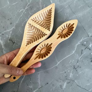 May include: Two wooden cookie moulds with carved designs. One mould features a leaf pattern, and the other has a sunburst design. The moulds are light brown and appear handmade. They are held against a grey marble surface.