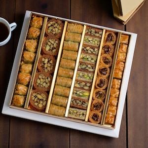 Könnte beinhalten: Eine Box mit verschiedenen Baklava-Gebäcksorten mit unterschiedlichen Formen und Füllungen, darunter Nüsse und Sirup. Das Gebäck ist in einer weißen Box mit goldenen Trennwänden angeordnet, neben einer Tasse Tee auf einer Holzoberfläche.