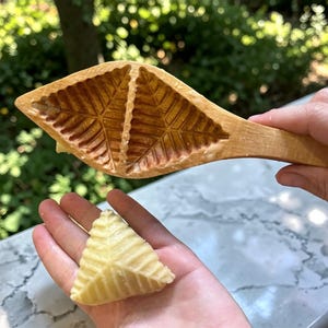 Puede incluir: Un molde de madera con un diseño de hojas se sostiene sobre una pasta triangular con un diseño de hojas a juego. El molde es de color marrón claro y la pasta es de color amarillo pálido. El fondo está borroso, mostrando vegetación.