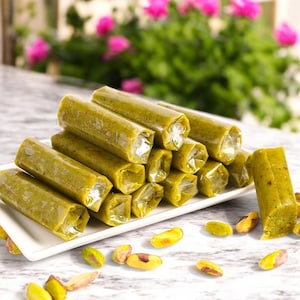 May include: A stack of green pistachio rolls wrapped in clear plastic on a white plate. The rolls are arranged in a pyramid shape. There are pistachio nuts scattered around the plate.