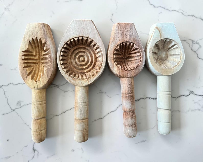 May include: Four wooden cookie molds with carved designs. The molds feature various patterns, including a sunburst, a floral design, and a spiral. Each mold has a handle for easy use. The wood has natural tones, with some molds painted white.