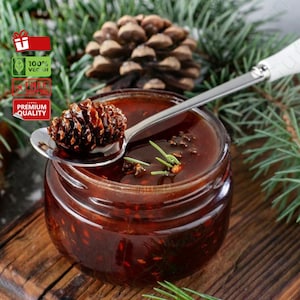 Puede incluir: Un tarro de vidrio lleno de un líquido espeso y viscoso de color rojo oscuro. Una cuchara con mango blanco sostiene un cono de pino cubierto con el mismo jarabe rojo. El tarro está sentado sobre una superficie de madera con ramas de pino en el fondo.