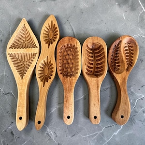 Puede incluir: Seis moldes de madera para galletas con varios diseños tallados, incluyendo helechos, rayos de sol y patrones geométricos. Cada molde tiene un mango y un agujero oscuro para colgar. Los moldes están hechos de madera clara.