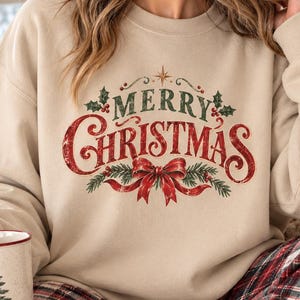 Peut inclure: Un sweat-shirt beige à col rond avec une impression graphique portant le texte Merry Christmas en lettres rouges et vertes, orné d'un nœud rouge, de branches de pin et de feuilles de houx. Une tasse blanche avec un sapin vert est tenue.
