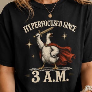 May include: A black short-sleeve t-shirt featuring a graphic of a white goose wearing a red cape and holding a sword. The text above the goose reads "HYPERFOCUSED SINCE" and the text below reads "3 A.M." with small star accents surrounding the goose.