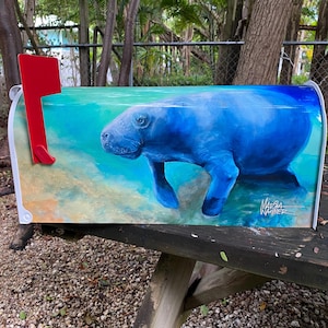 May include: A white mailbox with a red flag, featuring a vibrant painting of a blue manatee swimming underwater. The artwork includes shades of blue, green, and tan, with the artist's signature visible.