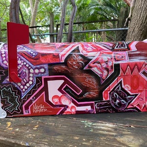 May include: A hand-painted mailbox with a vibrant, abstract design in shades of red, pink, and brown. The artwork includes geometric shapes, a cat silhouette, and the artist's signature. The mailbox has a red flag.