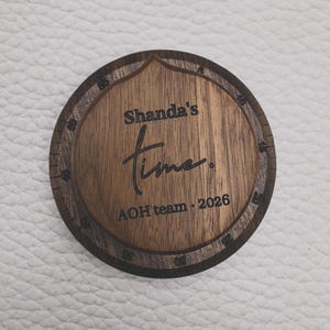 May include: A circular wooden plaque featuring a dark wood grain finish with a lighter center. The text "Shanda's time." is engraved in the middle, with "AOH team · 2026" below. The outer edge includes engraved numerical markings from 0 to 55.