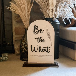 May include: A wooden sign with an arched top and a dark brown base. The sign reads "Be the Wheat" in black script, with a wheat stalk illustration. The text "LET BOTH GROW TOGETHER UNTIL HARVEST, MATTHEW 13" is printed at the bottom.