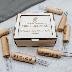May include: A wooden corn holder set in a square light-colored wooden box with a lid. The lid features an engraving of a corn cob and the text "CORN HOLDER SET For Family Cookouts & Summer Nights". Six wooden-handled corn picks with metal prongs.