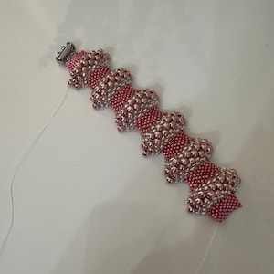 May include: A beaded bracelet with a pink and silver color scheme. The bracelet is made of small beads and has a decorative pattern. The clasp is silver. The bracelet is on a white surface.