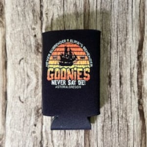 May include: Black neoprene can cooler with a vintage-style graphic of a ship sailing into a sunset. The text "Goonies Never Say Die!" is printed in white with a red outline. The words "Astoria, Oregon" are printed in white below the graphic.