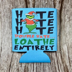 Könnte beinhalten: Ein blauer Getränkedosenkühler mit dem Text "HATE HATE HATE" in Schwarz und "DOUBLE HATE... LOATHE ENTIRELY" in Grün und Rot. Das Design zeigt eine grüne Cartoonfigur mit Weihnachtsmütze und rotem Hemd.
